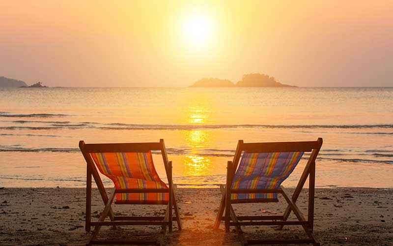 Two deck chairs sit on the beach, silhouetted against a vibrant sunset sky